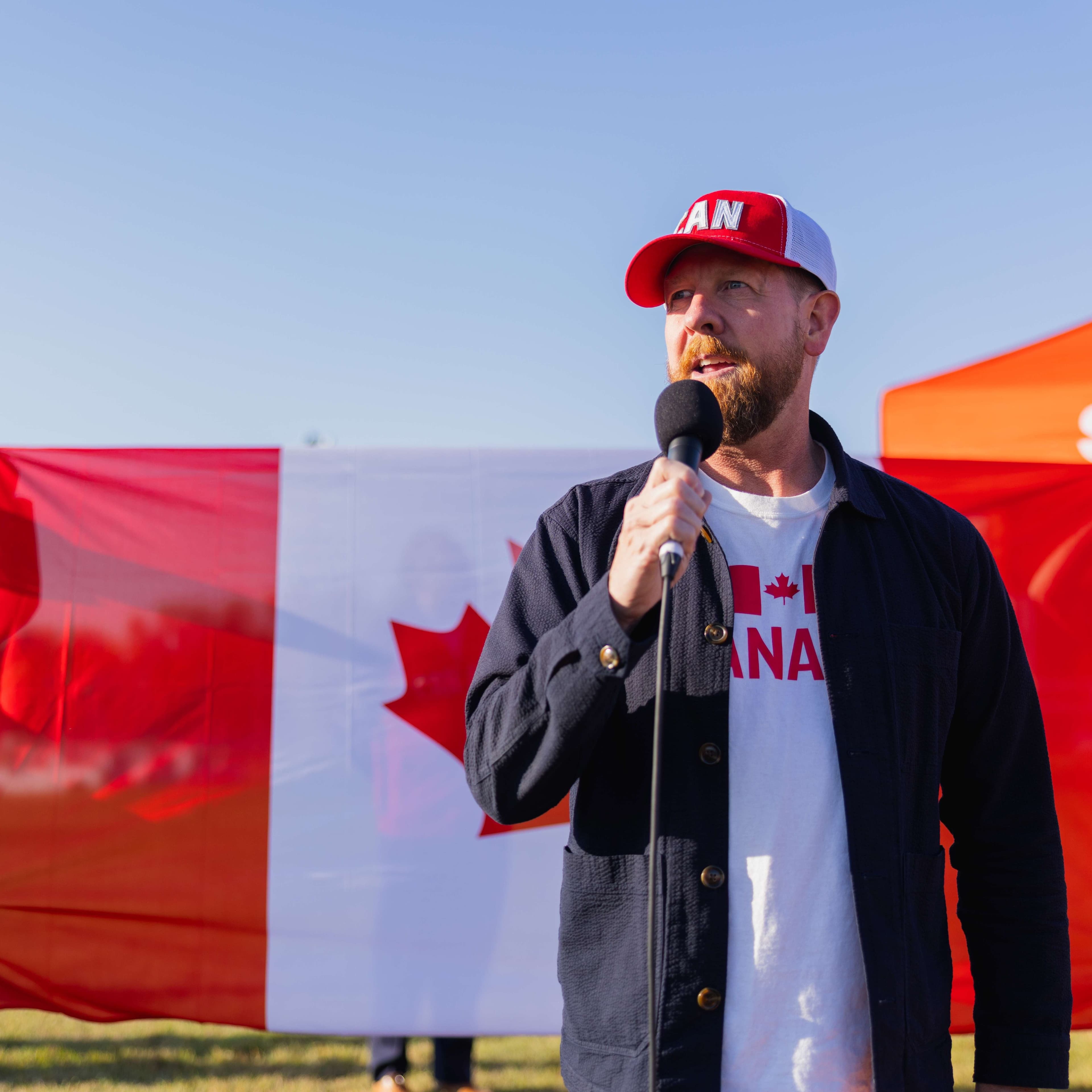MLA Kyle Kasawski holds a microphone while wearing a Canada Hat, dark jacket and Canada shirt.